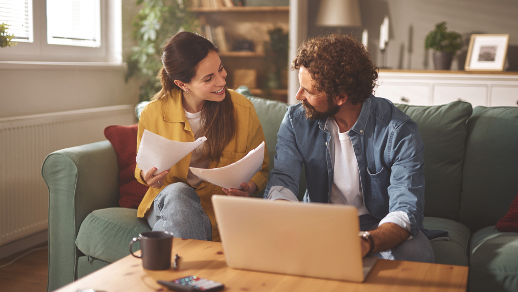 A couple sits together on their couch discussing various life insurance plans. They review important documents while a laptop is open on the coffee table, creating a warm atmosphere.