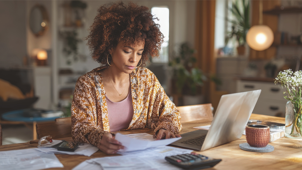 black woman accountant working remotely from home, sitting at dining table with open laptop and financial papers, focused on bookkeeping, auditing or reviewing tax returns, casual home clothes, calculator and coffee mug nearby, reading financial report for asset management and budgeting, cozy living room or kitchen background with natural daylight, realistic home office vibe, --ar 16:9 --v 7 Job ID: 13044516-0c38-4532-bb50-7b3a4bd562c4